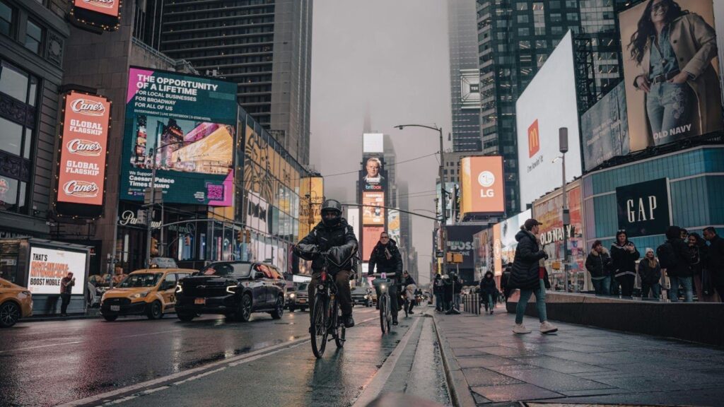 Cyclists ride through Times Square on a wet day in New York City, surrounded by taxis, cars, and digital billboards.