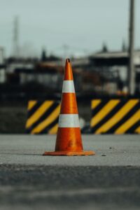 A single orange traffic cone placed on a roadway with blurred construction barriers in the background.