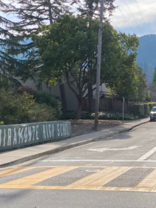 A street-level view of a crosswalk leading toward the entrance of Miramonte High School, with a green "Miramonte High School" banner on a chain-link fence.