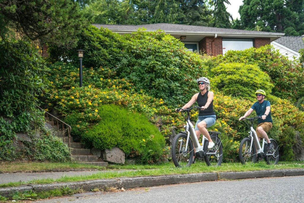 A man and a woman wearing helmets and sunglasses riding white electric bicycles on a paved suburban street past lush green landscaping and a brick house.