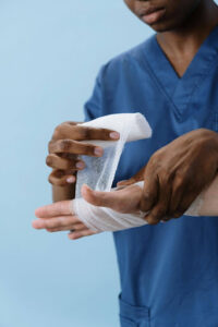 A healthcare provider wearing blue scrubs wrapping a white gauze bandage around a patient's injured wrist and hand.