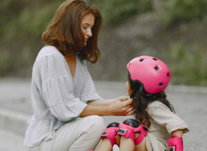 A mother kneeling outdoors to carefully fasten a bright pink bicycle helmet on her young daughter, who is also wearing pink knee and elbow pads.